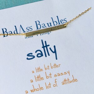 May include: A gold bar necklace with the text "BadAss Baubles and Things" on it. The necklace is on a white card with the text "satty" and the phrase "a little bit bitter, a little bit sassy, a whole lot of attitude" written in orange. The card also has the text "be Badass" at the bottom.