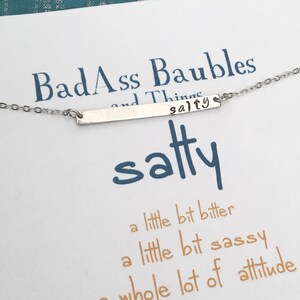 May include: A silver necklace with a bar pendant that says "salty" on it. The card below the necklace says "BadAss Baubles and Things" and "a little bit bitter, a little bit sassy, a whole lot of attitude".