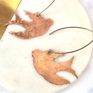 May include: A pair of copper-colored bird-shaped earrings with silver-colored ear wires. The earrings are displayed on a white and gold-colored circular surface. The birds have a textured appearance.