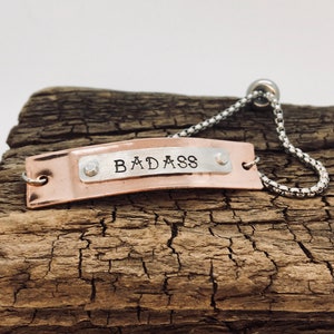 Könnte beinhalten: Ein Kupferarmband mit einer silbernen Kette und einer Kupferplatte, auf der "BADASS" in Großbuchstaben steht.