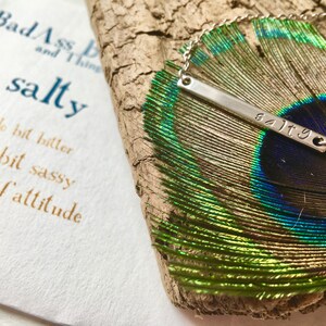 May include: A silver necklace with a bar pendant that says "salty" on a peacock feather. The necklace is on a wooden surface. The text "Bad Ass and Thing salty a little bit bitter little bit sassy a lot of attitude" is visible in the background.