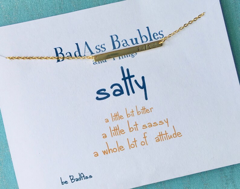 May include: A gold bar necklace with the text "BadAss Baubles and Things" on it. The necklace is on a white card with the text "satty" and the phrase "a little bit bitter, a little bit sassy, a whole lot of attitude" written in orange. The card also has the text "be Badass" at the bottom.