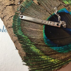 May include: A silver necklace with a bar pendant that says "salty" on a peacock feather background.
