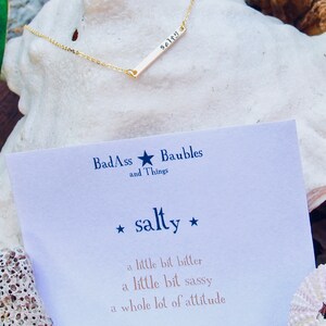 May include: A gold bar necklace with the word "salty" engraved on it. The necklace is displayed on a white seashell with a card that reads "BadAss and Things Baubles *salty* a little bit bitter a little bit sassy a whole lot of attitude"