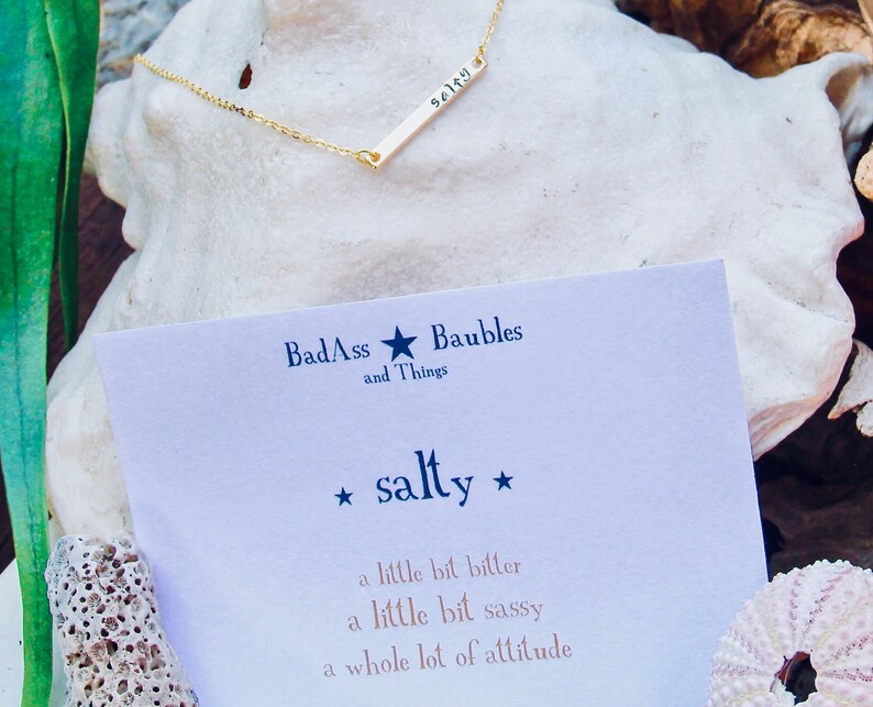 May include: A gold bar necklace with the word "salty" engraved on it. The necklace is displayed on a white seashell with a card that reads "BadAss and Things Baubles *salty* a little bit bitter a little bit sassy a whole lot of attitude"
