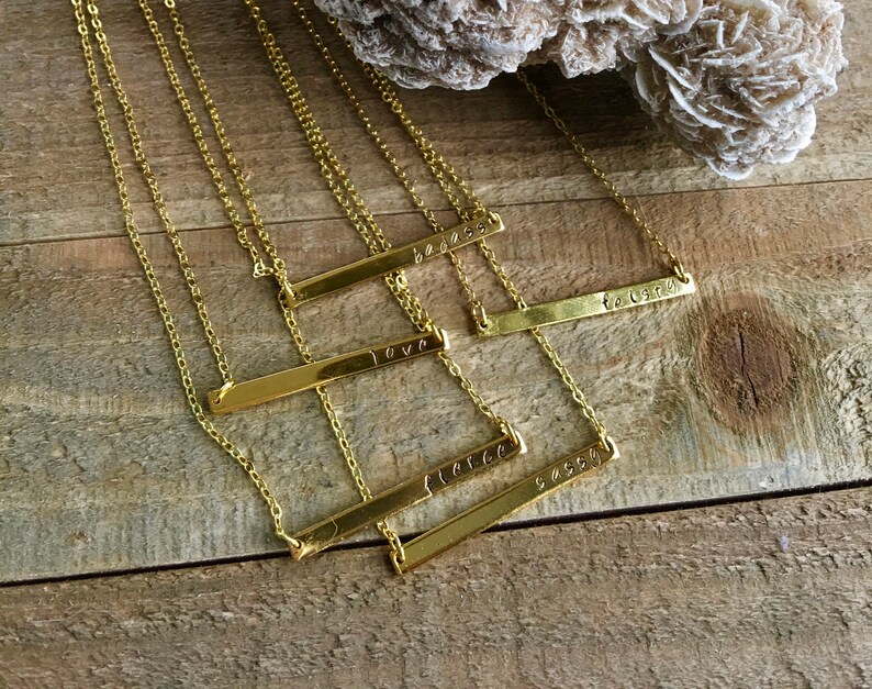 May include: Five gold bar necklaces with different words engraved on them. The words are "love", "be", "force", "sassy", and "together".