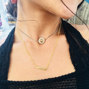 May include: A woman wearing a black shirt with two gold necklaces. The top necklace features a small, round pendant with a white flower design. The bottom necklace has a thin bar pendant with the word "goddess" engraved on it.