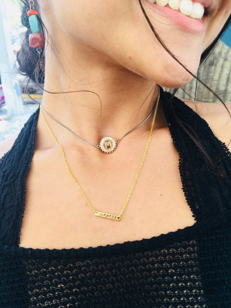 May include: A woman wearing a black shirt with two gold necklaces. The top necklace features a small, round pendant with a white flower design. The bottom necklace has a thin bar pendant with the word "goddess" engraved on it.