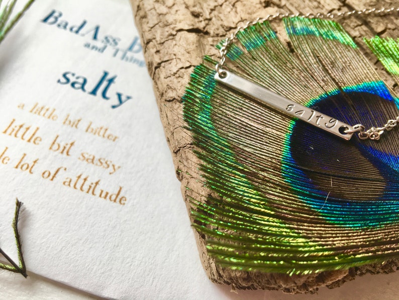 May include: A silver necklace with a bar pendant that says "salty" on a peacock feather. The necklace is on a wooden surface. The text "Bad Ass and Thing salty a little bit bitter little bit sassy a lot of attitude" is visible in the background.