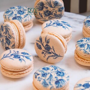 May include: A collection of pastel-colored macarons with intricate blue floral designs. The delicate cookies are arranged on a white surface, showcasing the detailed patterns and textures of the sweet treats. The image captures the elegance and artistry of the baked goods.