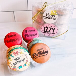 May include: A clear plastic box filled with colorful macarons. The macarons are decorated with the words "Mama Philippa", "Happy Birthday", and "IZZY macarons".