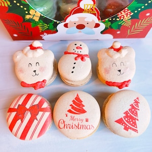 May include: Six Christmas-themed macarons. Two are white with red accents and bear faces, one is white with black accents and a snowman face, one is white with red and white stripes and a red bow, one is white with red accents and a Christmas tree, and one is white with red accents and a Christmas tree with white dots.