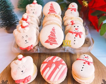 May include: A collection of Christmas-themed macarons in a clear plastic container. The macarons are decorated with images of snowmen, Christmas trees, and polar bears. The text "IZZY macarons" is visible on the container.