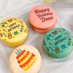 May include: Four colorful macarons with birthday greetings. One macaron is pink with the text "Happy Birthday Dana". Another macaron is yellow with the text "Happy Birthday!" and colorful birthday-themed illustrations. A third macaron is green with the text "Happy Birthday!" and colorful birthday-themed illustrations. The fourth macaron is beige with a colorful illustration of a birthday cake.