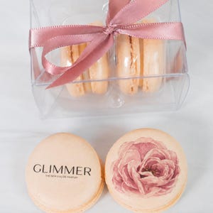 May include: A clear plastic box tied with a dusty rose ribbon containing light peach macarons. Two additional macarons sit in front: one features the black text GLIMMER THE NEW EAU DE PARFUM, and the other displays a printed illustration of a pink rose.