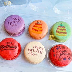 May include: Six colorful macarons in a clear plastic container. The macarons are decorated with birthday themes, including the words "Happy Birthday Alice" and a cake with candles.