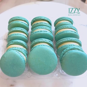 May include: A clear plastic container holds a dozen turquoise macarons with a pale yellow filling. The macarons are arranged in three rows of four. The text "IZZY MACARONS" is in the upper right corner.
