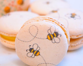 May include: Close-up of several light-colored macarons, some with bee illustrations. The macarons have a light orange filling. The text "Izzy Macarons" is visible in the background. The bee design includes yellow and black details.