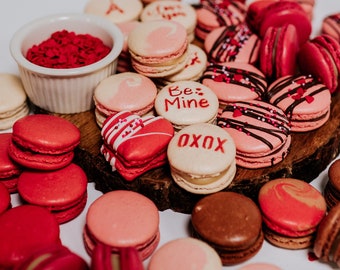May include: A variety of pink, red, and brown macarons with different designs and decorations. Some macarons have messages like "Be Mine" and "XOXO" written on them.