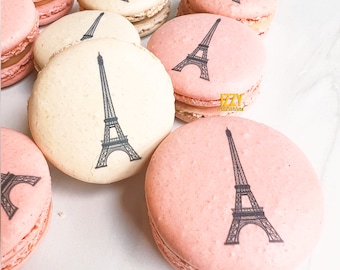 May include: A collection of round macarons in shades of pale pink and cream, each featuring a black printed graphic of the Eiffel Tower. The text IZZY macarons is printed in yellow on one of the pink cookies, showcasing a delicate texture.