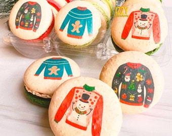 Può includere: Sei macarons a tema natalizio con diversi design. I macarons sono disposti in un contenitore di plastica trasparente. I macarons sono decorati con immagini di maglioni con motivi natalizi, tra cui un pupazzo di neve, un uomo di pan di zenzero e Babbo Natale.