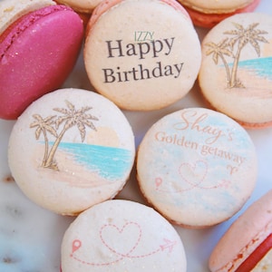 May include: A collection of colorful macarons with various designs. Some macarons feature beach scenes with palm trees and ocean, while others have text that reads "Happy Birthday" and "Shay's Golden getaway".