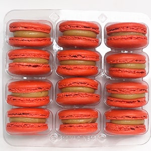 May include: A clear plastic tray holds a dozen vibrant red macarons. Each macaron has a light brown filling, creating a two-toned effect. The cookies are arranged in three rows of four, showcasing a classic French pastry.