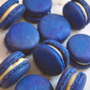 May include: Close-up of several blue macarons with a creamy, pale yellow filling. The macarons are arranged on a white surface. The shells have a marbled effect, and the filling is visible between the two halves of each macaron.