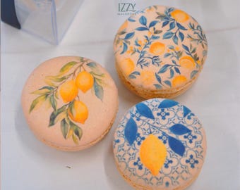 Può includere: Tre macaron rotondi color beige con disegni stampati. Uno presenta un ramo di limone con frutti gialli e foglie verdi. Un altro mostra un motivo di piccoli limoni gialli e fogliame blu. Il terzo mostra un singolo limone giallo su un motivo a piastrelle.