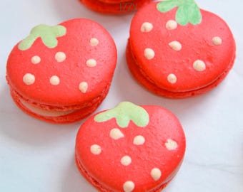 May include: Four strawberry-shaped macarons. Each macaron is bright red with white dots and a green stem. The macarons are arranged on a white surface.