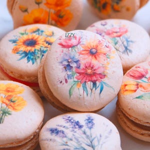 May include: Close-up of several macarons with floral designs. Each macaron features a different watercolor flower arrangement in shades of pink, yellow, blue, and purple. The macarons are light brown with a cream filling. The logo "IZZY MACARONS" is visible.