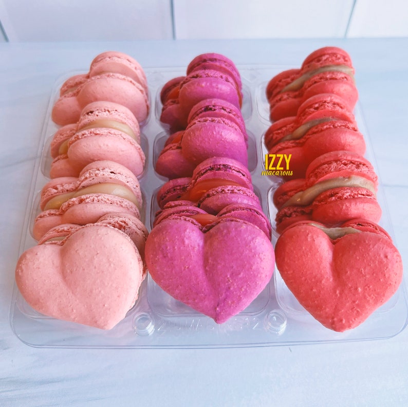 May include: A clear plastic container filled with heart-shaped macarons. The macarons are arranged in three rows of four. The macarons are pink, red, and white. The container has the text "IZZY macarons" printed on it.