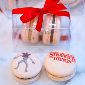 May include: A clear plastic box tied with a red ribbon contains two macarons. Two additional macarons are in front of the box. One macaron has a Demogorgon graphic, and the other has the text "STRANGER THINGS" in red.