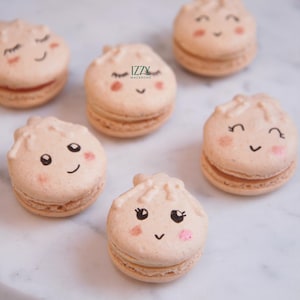 May include: Several light brown macarons with adorable dumpling-like faces, featuring black eyes, rosy cheeks, and smiling mouths. The macarons are arranged on a white surface, with the "Izzy Macarons" logo visible on one.
