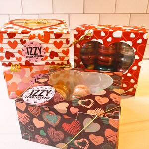 May include: Three gift boxes with heart-shaped designs. The boxes are filled with macarons. The boxes are decorated with the text "IZZY macarons".