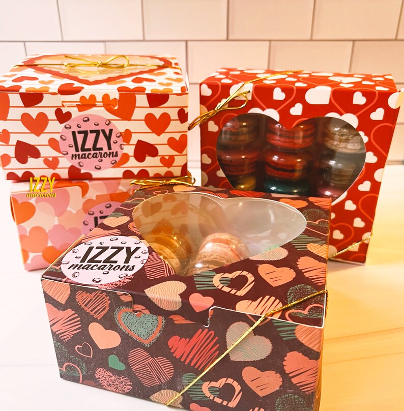 May include: Three gift boxes with heart-shaped designs. The boxes are filled with macarons. The boxes are decorated with the text "IZZY macarons".