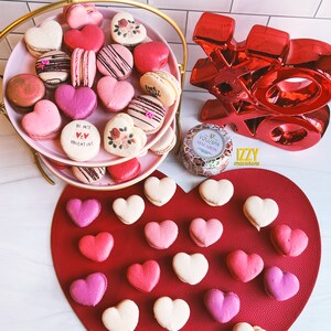 May include: A two-tiered gold stand with pink heart-shaped macarons decorated with various designs, including flowers, hearts, and the phrase "Be My Valentine". The stand is on a white surface with a red heart-shaped mat with additional pink heart-shaped macarons. A red "XO" sign and a candle are also visible.