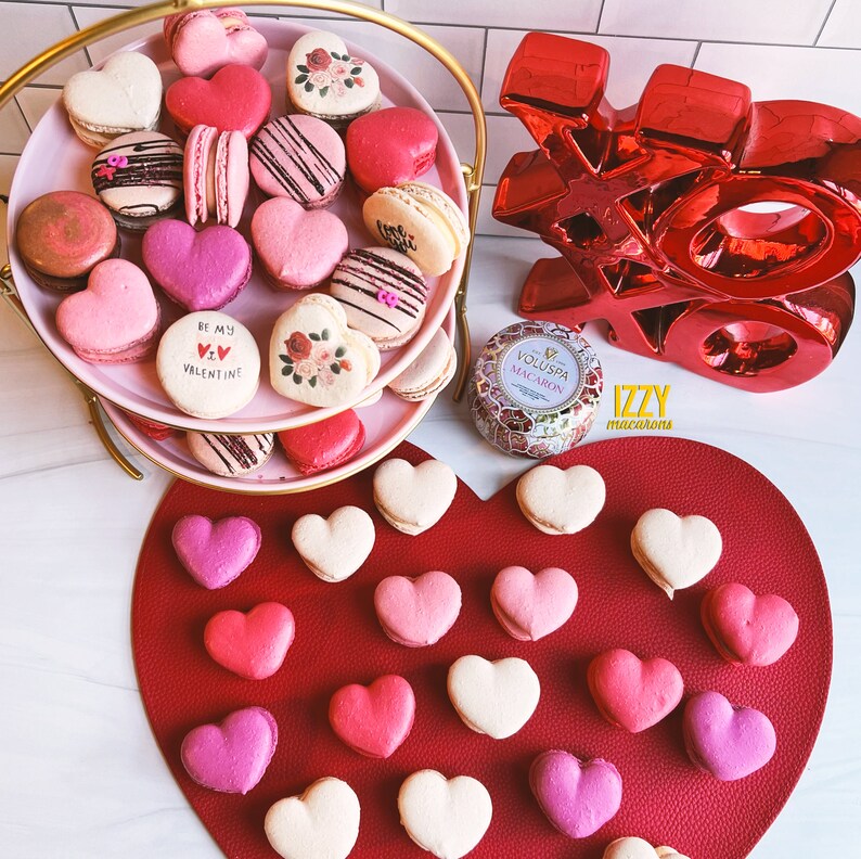 May include: A two-tiered gold stand with pink heart-shaped macarons decorated with various designs, including flowers, hearts, and the phrase "Be My Valentine". The stand is on a white surface with a red heart-shaped mat with additional pink heart-shaped macarons. A red "XO" sign and a candle are also visible.