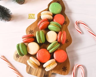 May include: A wooden, Christmas tree-shaped serving board filled with colorful macarons in shades of red, green, and cream. Candy canes and small green Christmas trees surround the board. The text "IZZY macarons" is visible.