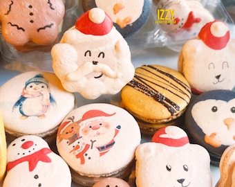 May include: Assortment of Christmas-themed macarons with various designs, including a penguin, snowman, Santa Claus, gingerbread man, and a polar bear. The macarons are arranged in a clear plastic container with the text "IZZY macarons" printed on the side.