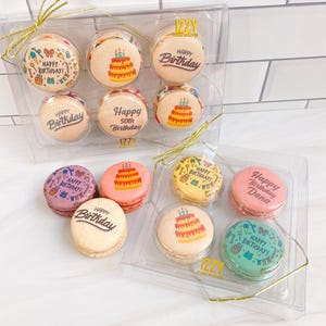 May include: Assortment of colorful macarons with various birthday messages. The macarons are arranged in two clear plastic boxes. The macarons are decorated with colorful frosting and sprinkles. The boxes are labeled "IZZY macarons".