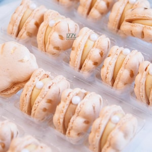 May include: Close-up of a clear plastic container filled with several light-colored macarons. Each macaron has a pale yellow filling and a small white pearl-like decoration. The macarons are arranged in rows. The text "IZZY MACARONS" is visible.