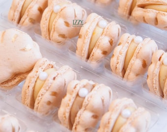 May include: Close-up of a clear plastic container filled with several light-colored macarons. Each macaron has a pale yellow filling and a small white pearl-like decoration. The macarons are arranged in rows. The text "IZZY MACARONS" is visible.