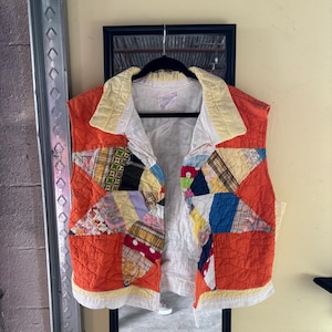 Puede incluir: Un chaleco acolchado con un diseño de patchwork. El chaleco presenta una base naranja vibrante con parches de tela coloridos en varios patrones. Tiene un cuello amarillo y forro blanco. El chaleco está colgado en una percha negra.