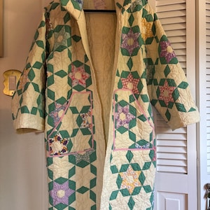 May include: A long-sleeved coat constructed from a cream-colored quilt featuring a repeating star pattern in shades of green, purple, yellow, and pink. The garment includes two large front pockets outlined with pink trim and a high collar.