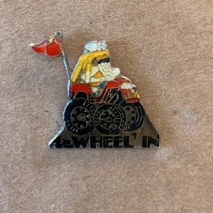Puede incluir: Un pin de plata con una caricatura de una persona conduciendo un camión monstruo rojo con ruedas negras. El camión está subiendo una colina con las palabras "Wheel'in" en el lateral.