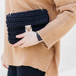 May include: A black crocheted shoulder bag with a gold clasp and a gold chain strap. The bag is being held by a person wearing a brown sweater.