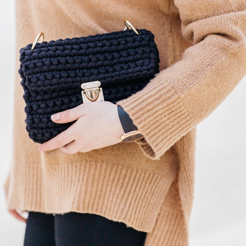 May include: A black crocheted shoulder bag with a gold clasp and a gold chain strap. The bag is being held by a person wearing a brown sweater.