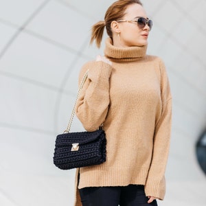 May include: A woman wearing a tan turtleneck sweater and black pants is carrying a black crocheted handbag with a gold chain strap.