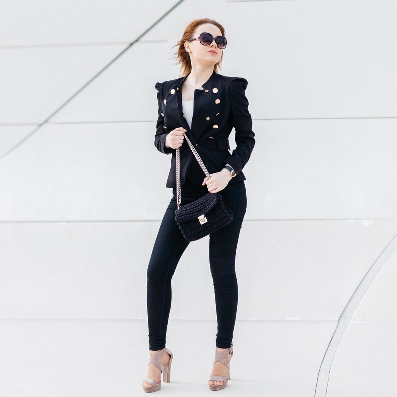 May include: A woman wearing a black blazer with gold buttons, black pants, and black heeled sandals. She is carrying a black chain strap bag.